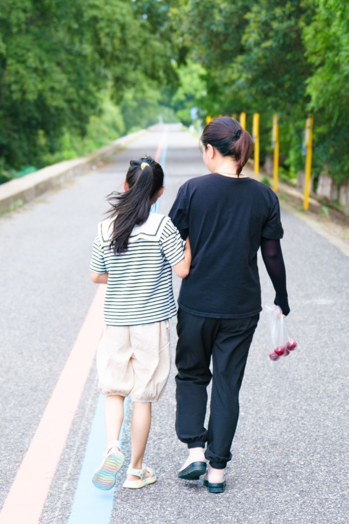 A mother and daughter walking together on a tree-lined road during the day.